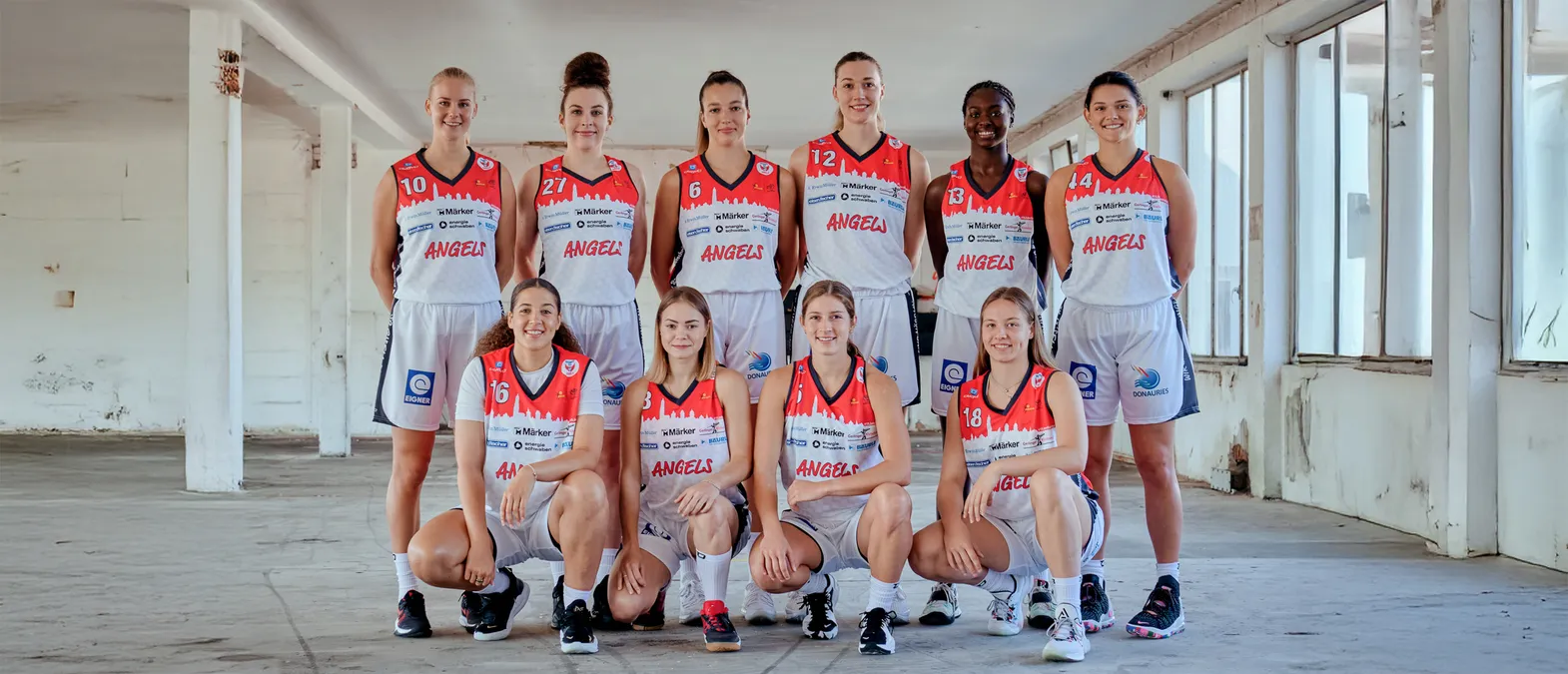 A group of eight women in orange and white basketball jerseys with the word 'ANGELS' on them pose in a warehouse.
