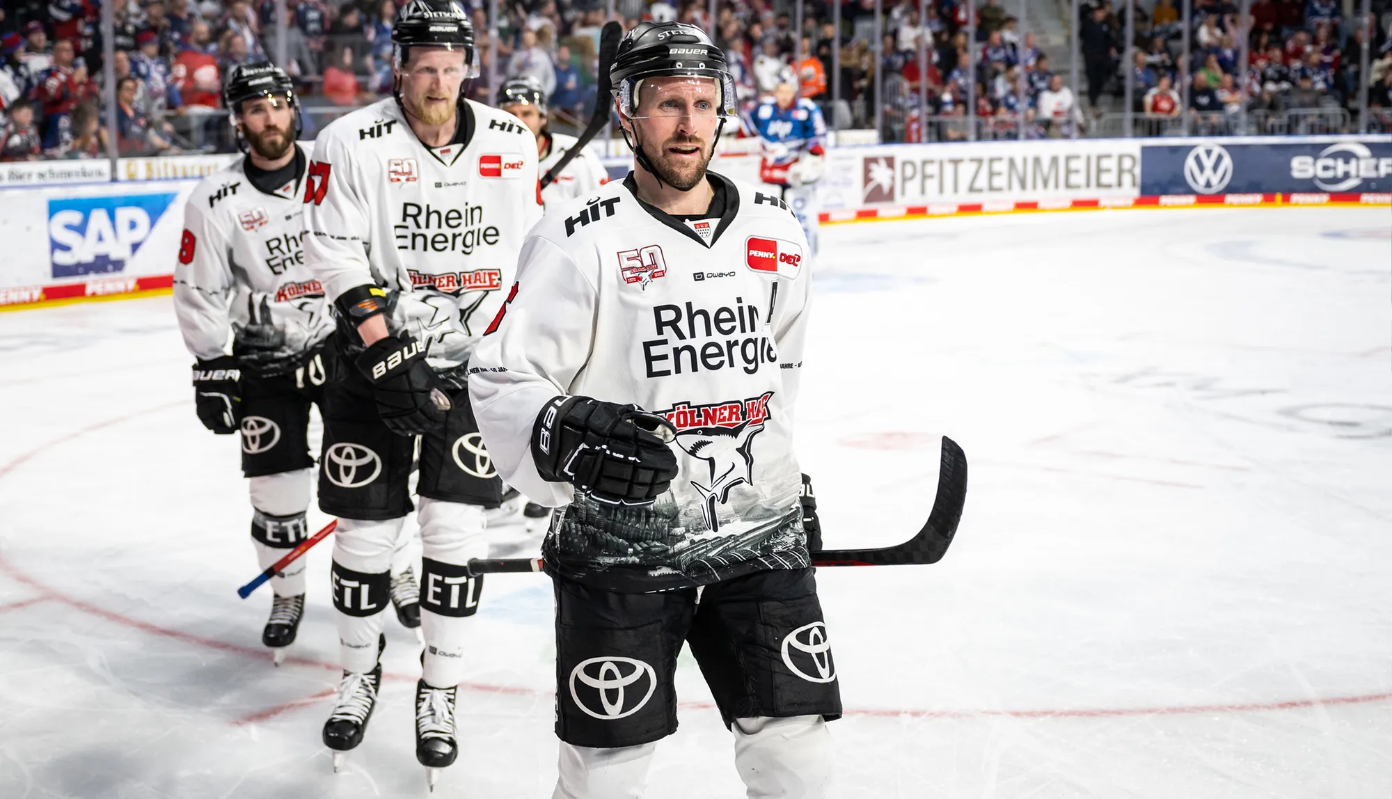 Three ice hockey players in white jerseys with different numbers stand on the ice during a game.