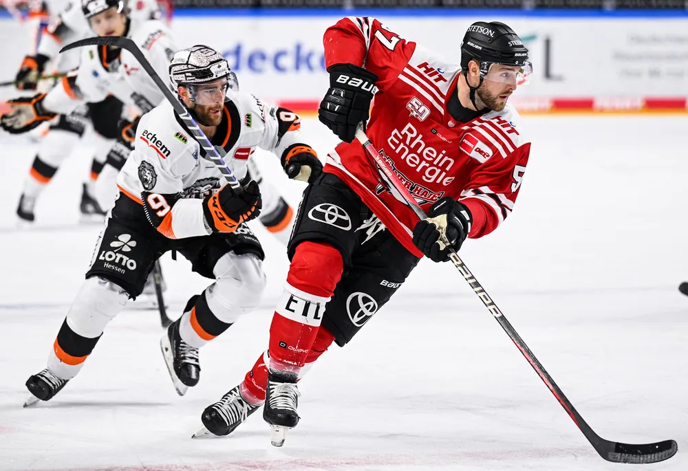 A hockey player in a red jersey skates with the puck on the ice while another player in a white jersey follows closely behind.