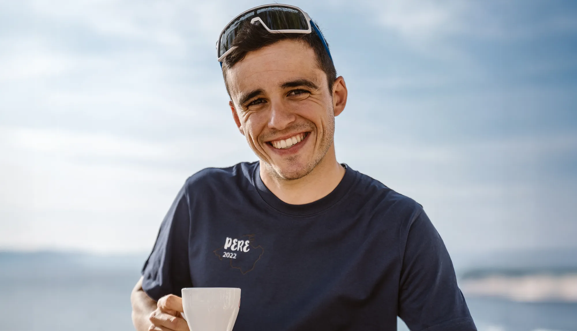 A young man smiles in a blue T-shirt featuring the text 'PERE 2022' while holding a cup, set against a clear sky.