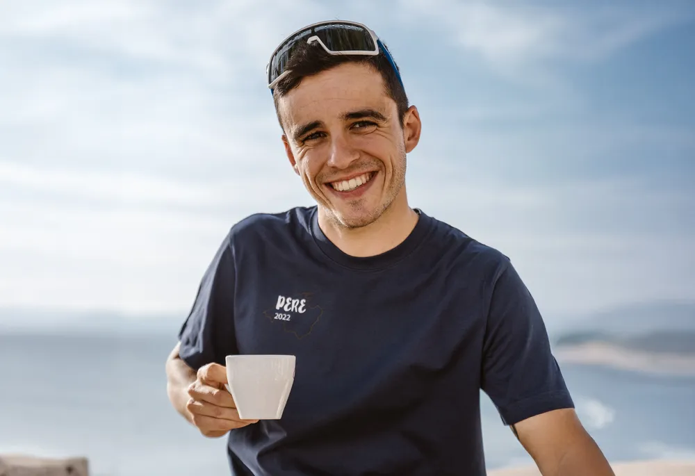 A smiling man in a blue t-shirt holds a cup in his hand, set against a beautiful coastal landscape.