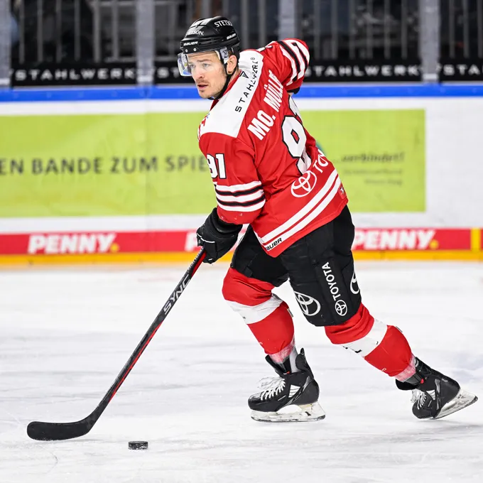 A hockey player in a red and black jersey with the number 91 and the name Mc. Miller on the back, standing on the ice and controlling a puck with his stick.