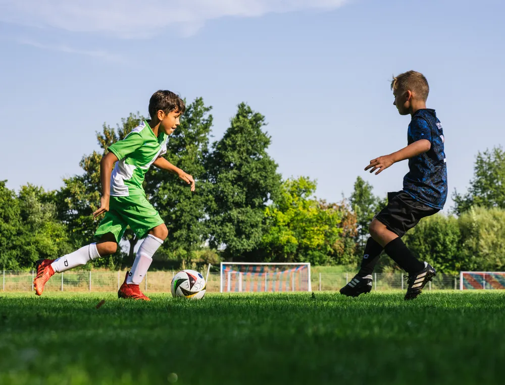 Two children are playing soccer on a green field, one wearing a green jersey and the other in black, as they chase the ball.