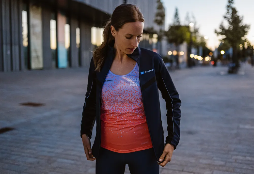 A sporty woman is wearing a dark blue jacket over a gradient sleeveless top in blue and orange while standing in an urban environment.