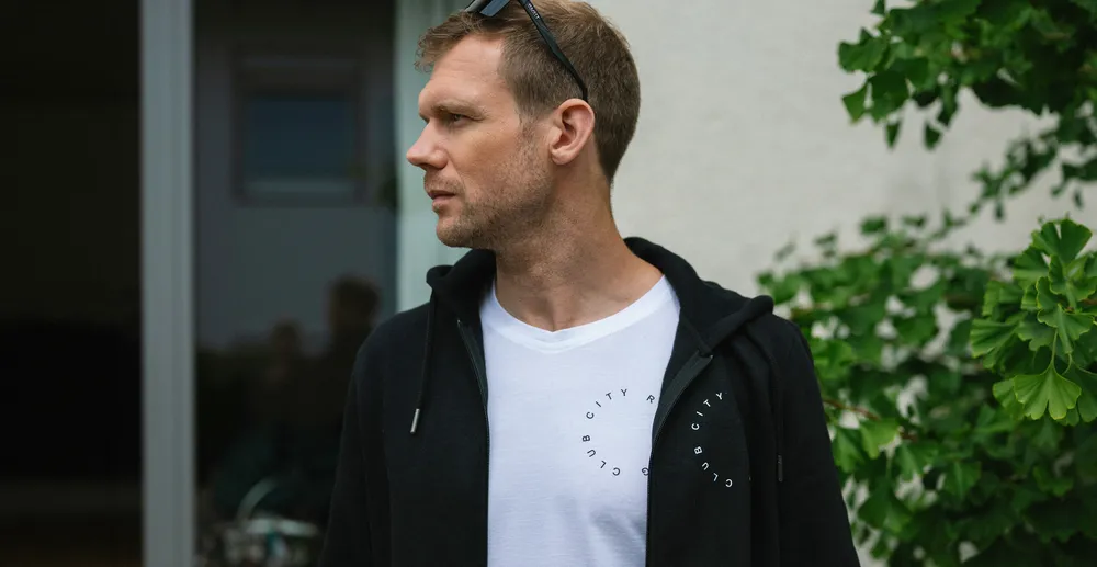 A man stands sideways wearing a black hoodie over a white T-shirt with a print, surrounded by plants.