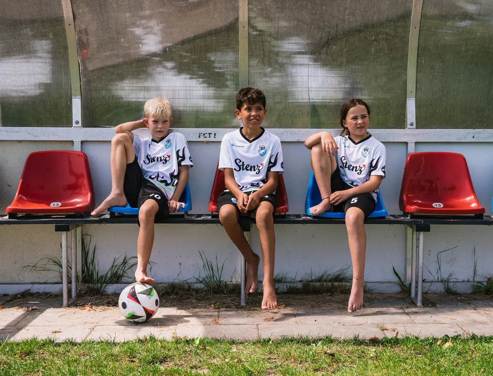 Three children sit on a bench at the edge of a soccer field, wearing custom-designed soccer jerseys with a soccer ball in front of them.