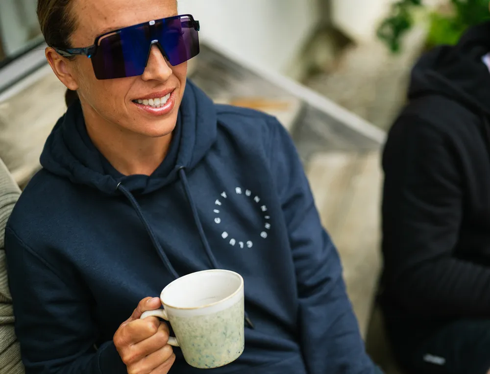 A person in a navy hoodie with a logo holds a coffee cup and wears large sunglasses.