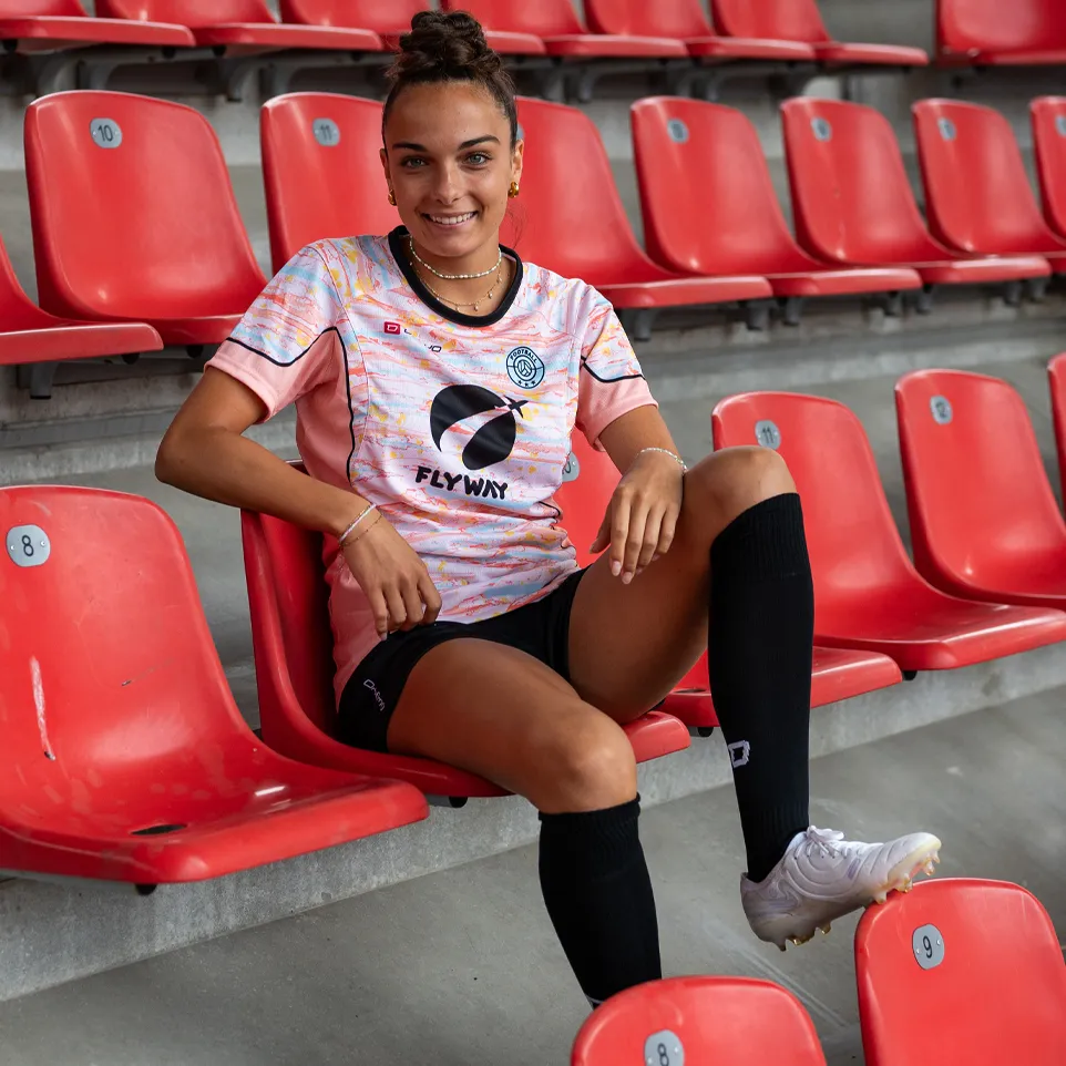 A smiling woman sits in a stadium on a red seat, wearing a colorful soccer jersey and black socks.