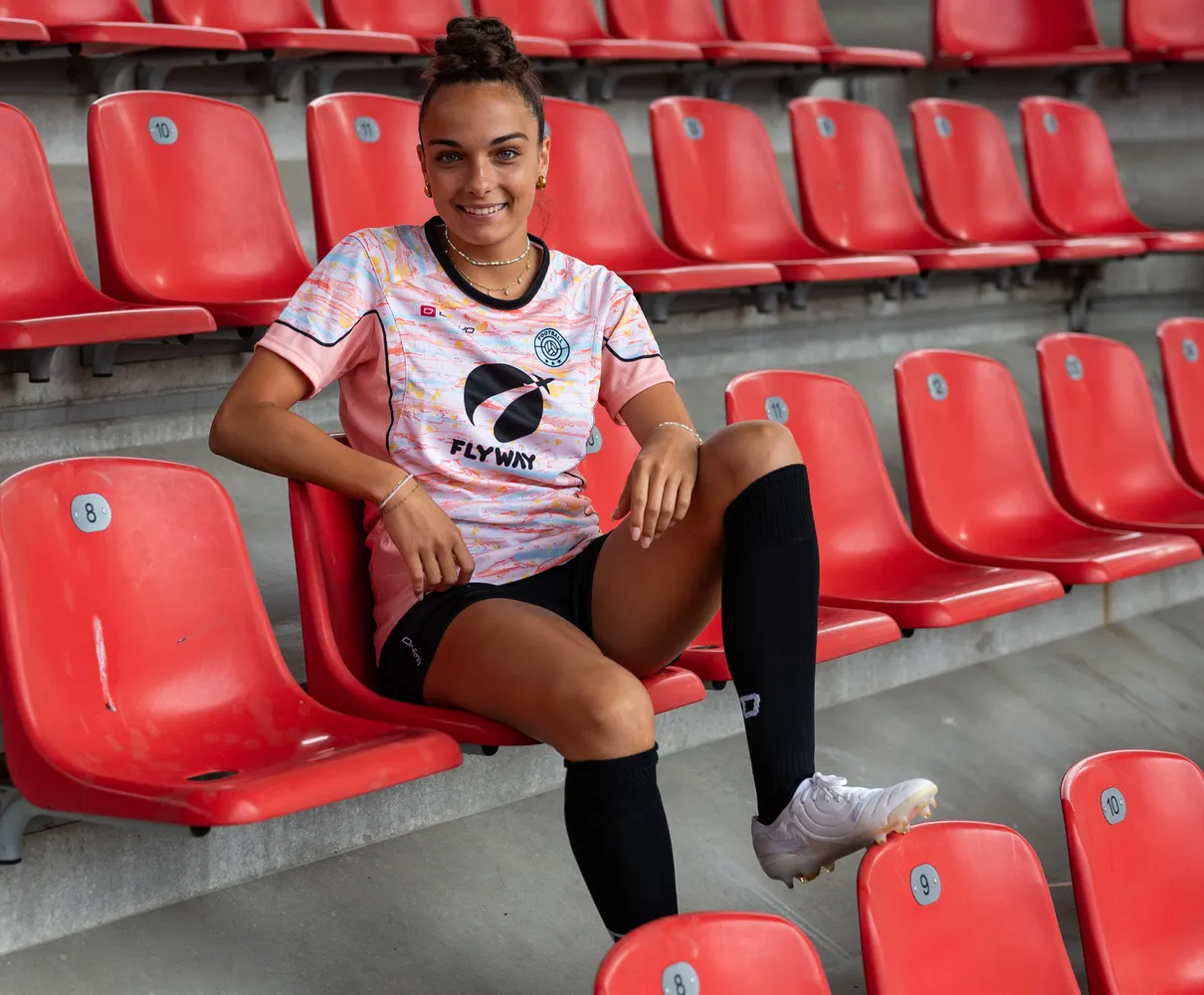 A young woman sits on a red stadium seat wearing a colorful soccer jersey and black socks.