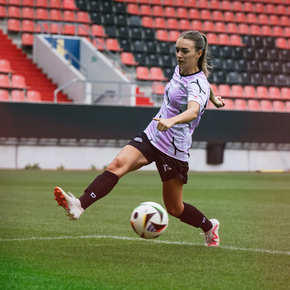A female player is kicking a soccer ball on a stadium field, wearing a purple jersey and black shorts.