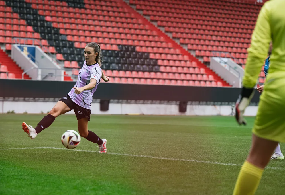 A female soccer player in a purple and black jersey kicks a ball on a field with empty stands in the background.
