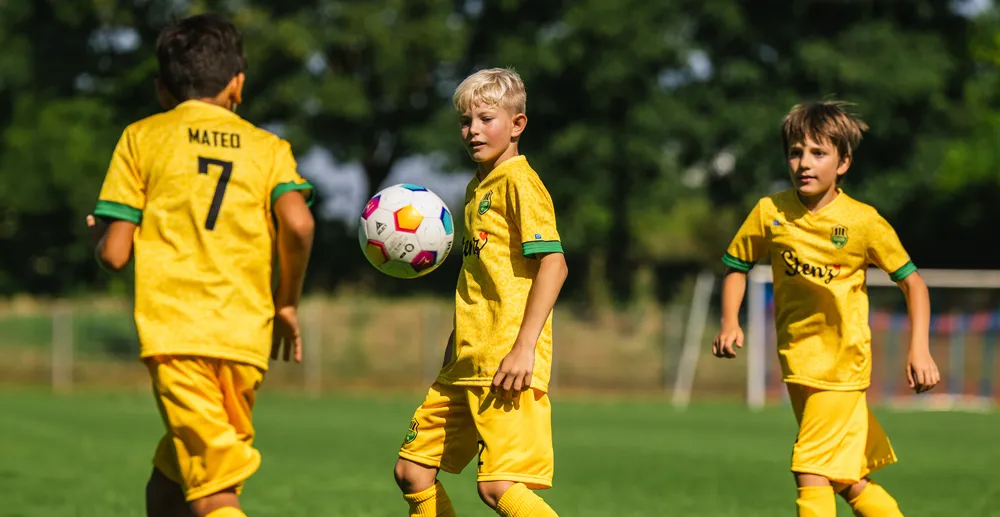 Three children in yellow soccer jerseys are playing on a green field, with one of them controlling a colorful soccer ball.
