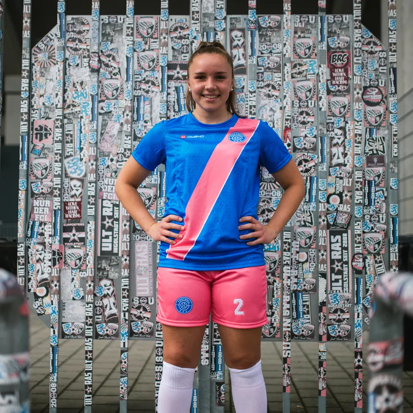 A young girl in a blue soccer jersey with a pink stripe and pink shorts stands in front of a wall covered in stickers.