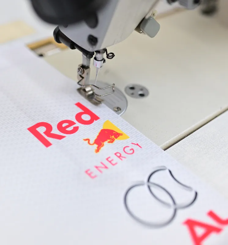 A sewing machine is stitching a white sports garment with a red Red Bull logo.