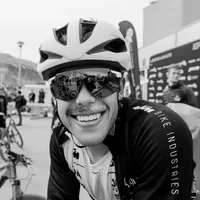 A smiling cyclist wearing a black cycling jacket with 'BIKE INDUSTRIES' printed on the sleeve and a white helmet, wearing sunglasses reflecting the surroundings.