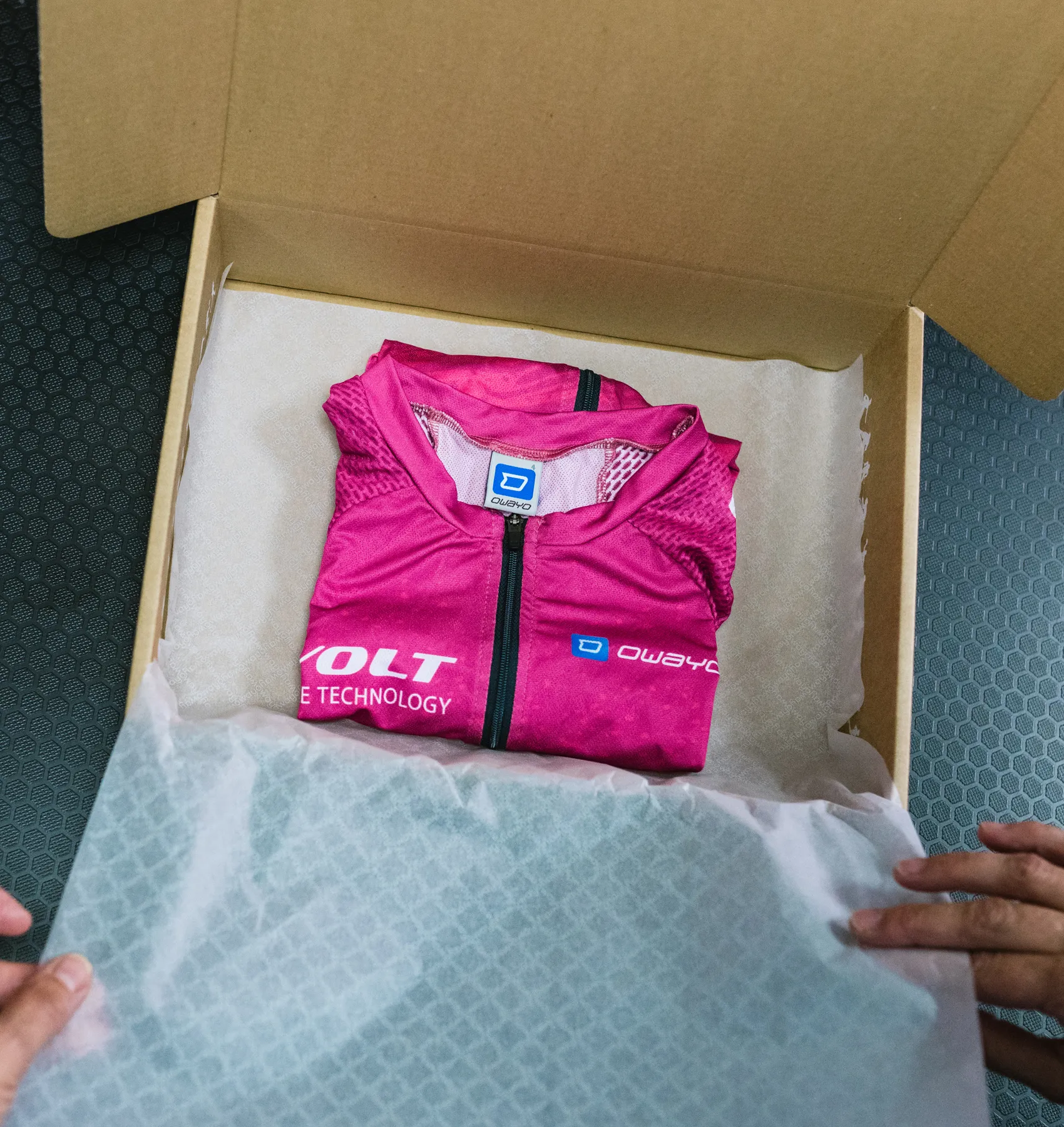 A folded, pink, customized sportswear piece from owayo is nestled in a cardboard box lined with white paper, being held by hands.