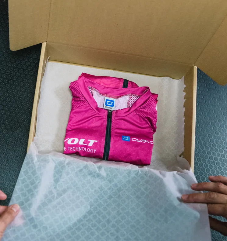A folded, pink, customized sportswear piece from owayo is nestled in a cardboard box lined with white paper, being held by hands.