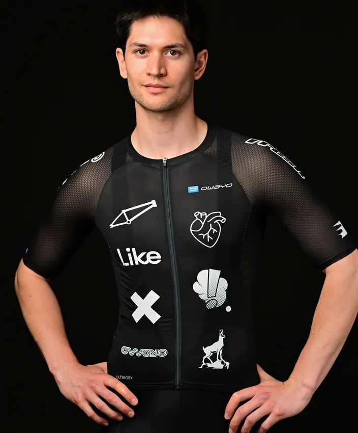 A young man is wearing a fitted black sports top adorned with various white logos and designs, posing against a black background.