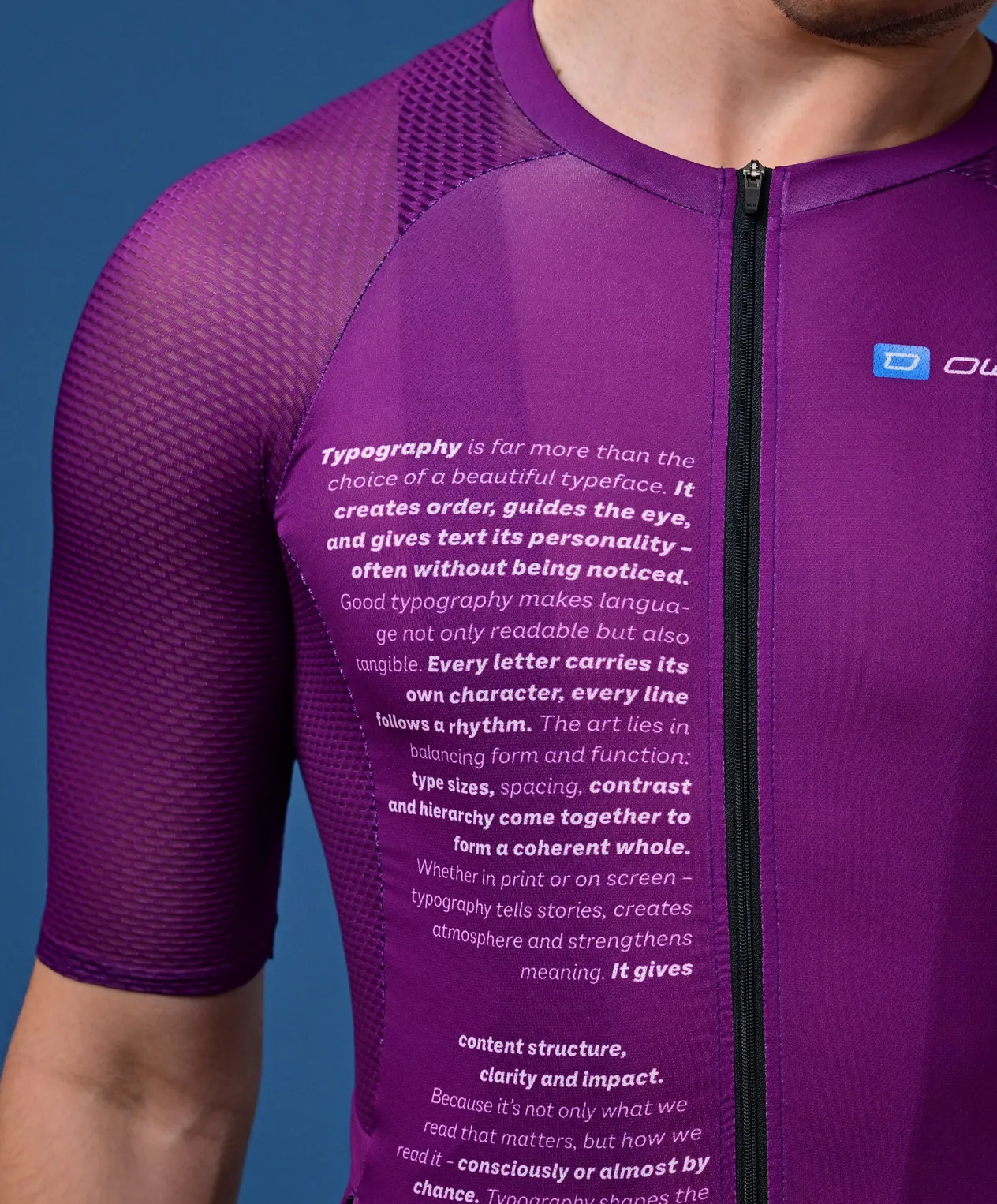 A man wears a purple, short-sleeved sports jersey with a text about typography on the chest and a small logo on the shoulder.