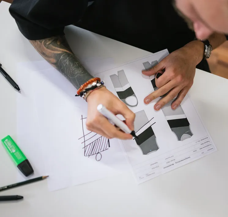 A person with a tattooed arm sketches designs for customized sportswear with a marker on a sheet of paper.