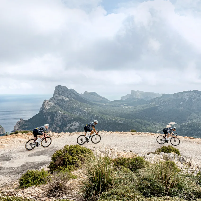 Three cyclists in athletic wear ride along a narrow path with a view of a coastal landscape.