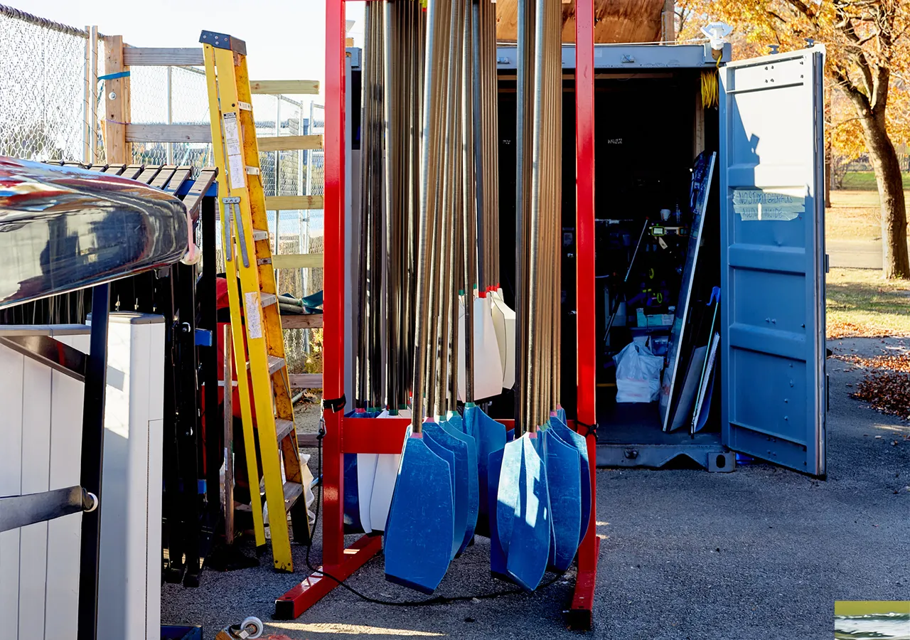 An image of a boat storage area featuring oars in various colors, a yellow ladder, and an open container in the background.