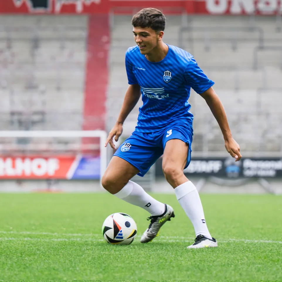 A young football player in a blue jersey and blue shorts with a logo on the chest and the number 3 on the shorts dribbles a football on a green grass field.
