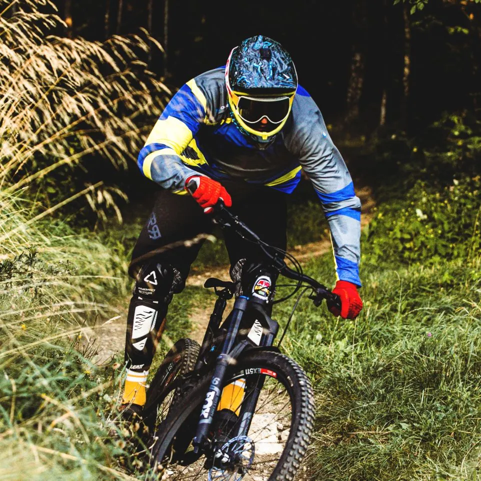A mountain biker wearing a colorful long-sleeved jersey with blue, yellow, and gray accents rides on a forest trail.