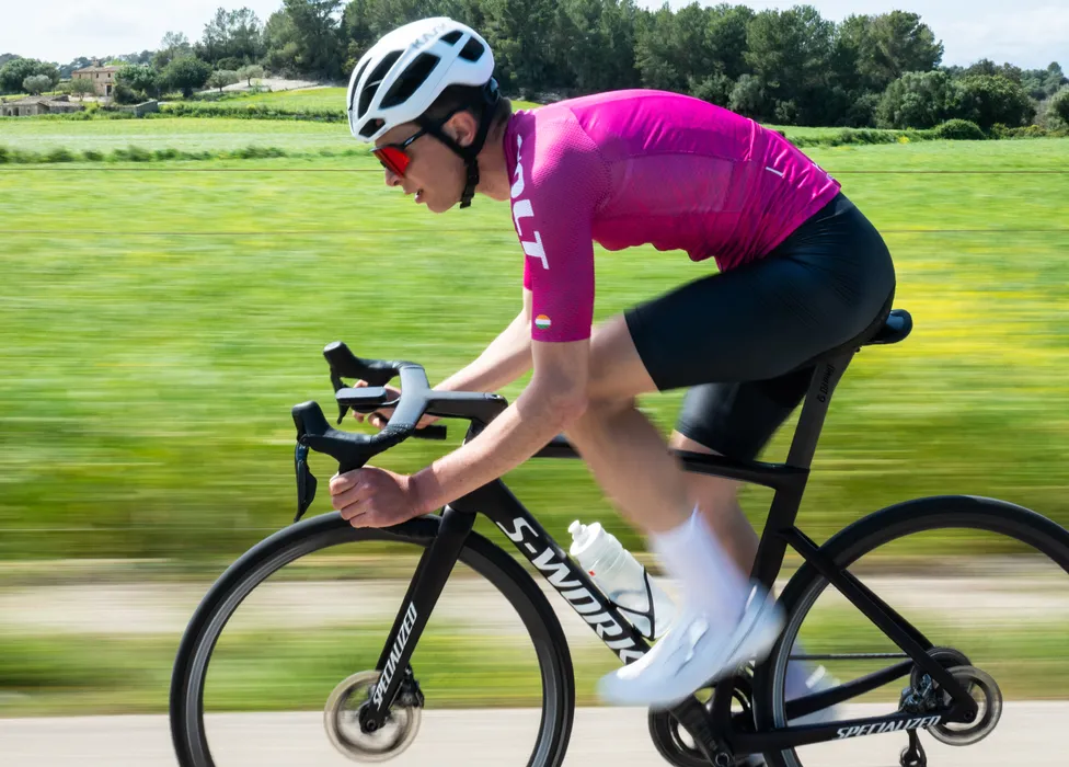 A cyclist in a pink jersey and black shorts rides a road bike on a country road, with a blurred green background suggesting speed.